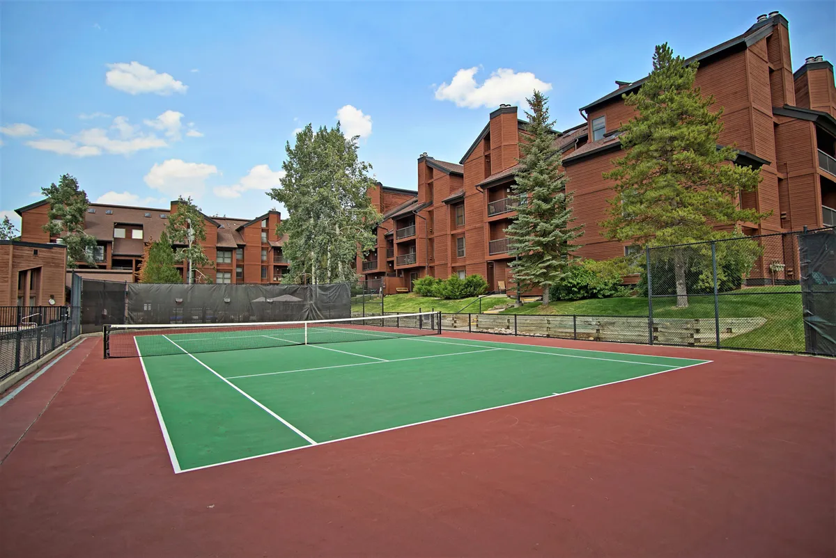 An on-site tennis court framed by greenery and residential buildings, capturing an active, social atmosphere. The scene invites guests to picture themselves engaging in friendly summer matches or enjoying community events outdoors.