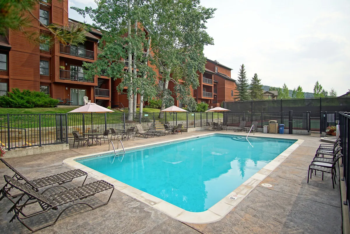 A heated outdoor swimming pool surrounded by lounge chairs and umbrellas, with lush green trees and a building in the background. The serene atmosphere suggests relaxation and leisure, perfect for enjoying the summer sun. The presence of amenities like seating areas supports group socializing and poolside gatherings.