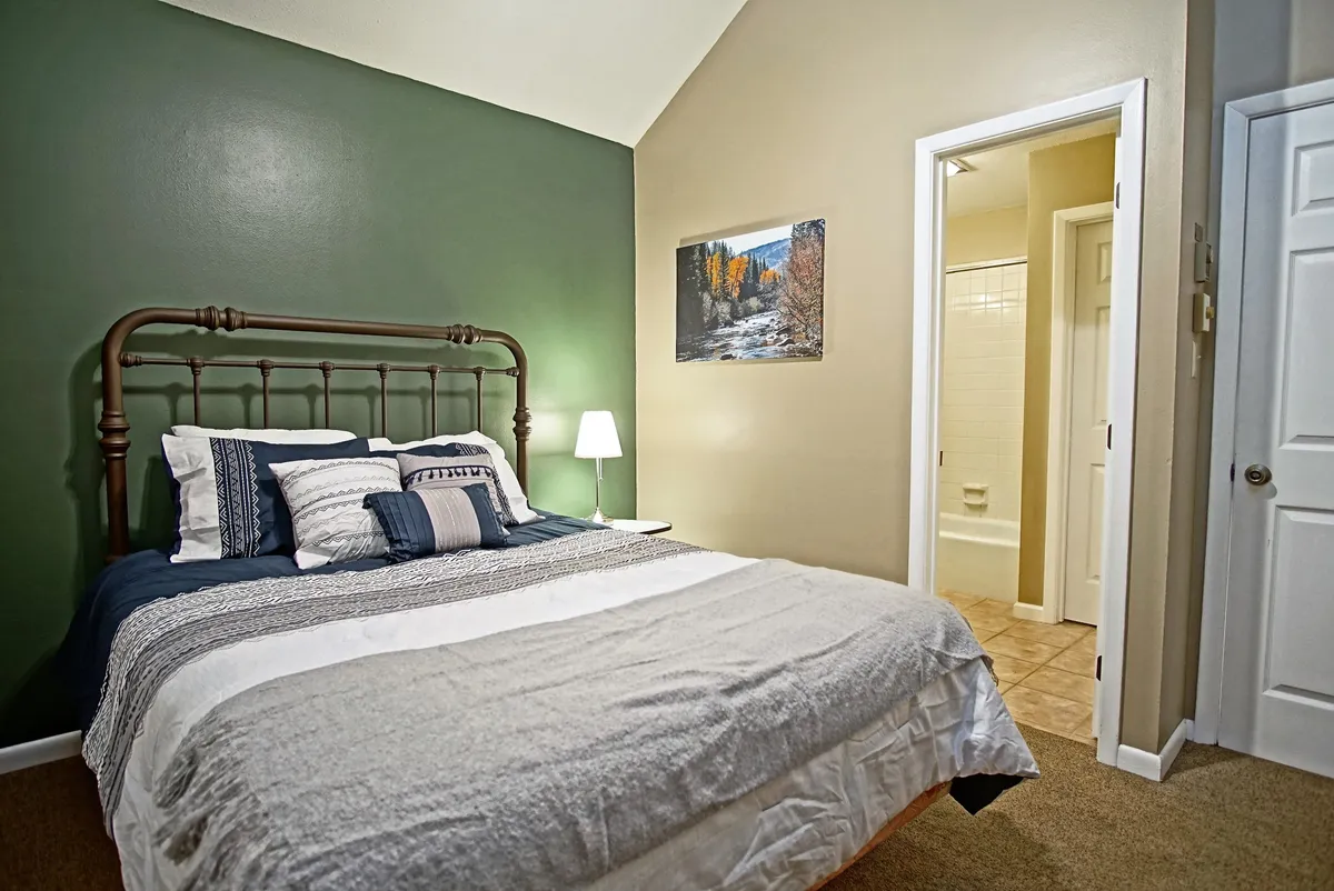 A tranquil bedroom with a large bed adorned with decorative pillows, a soft-lit lamp on the nightstand, tasteful wall art, and a window allowing for gentle, natural light. The space exudes peace and is perfect for couples seeking a calm retreat.