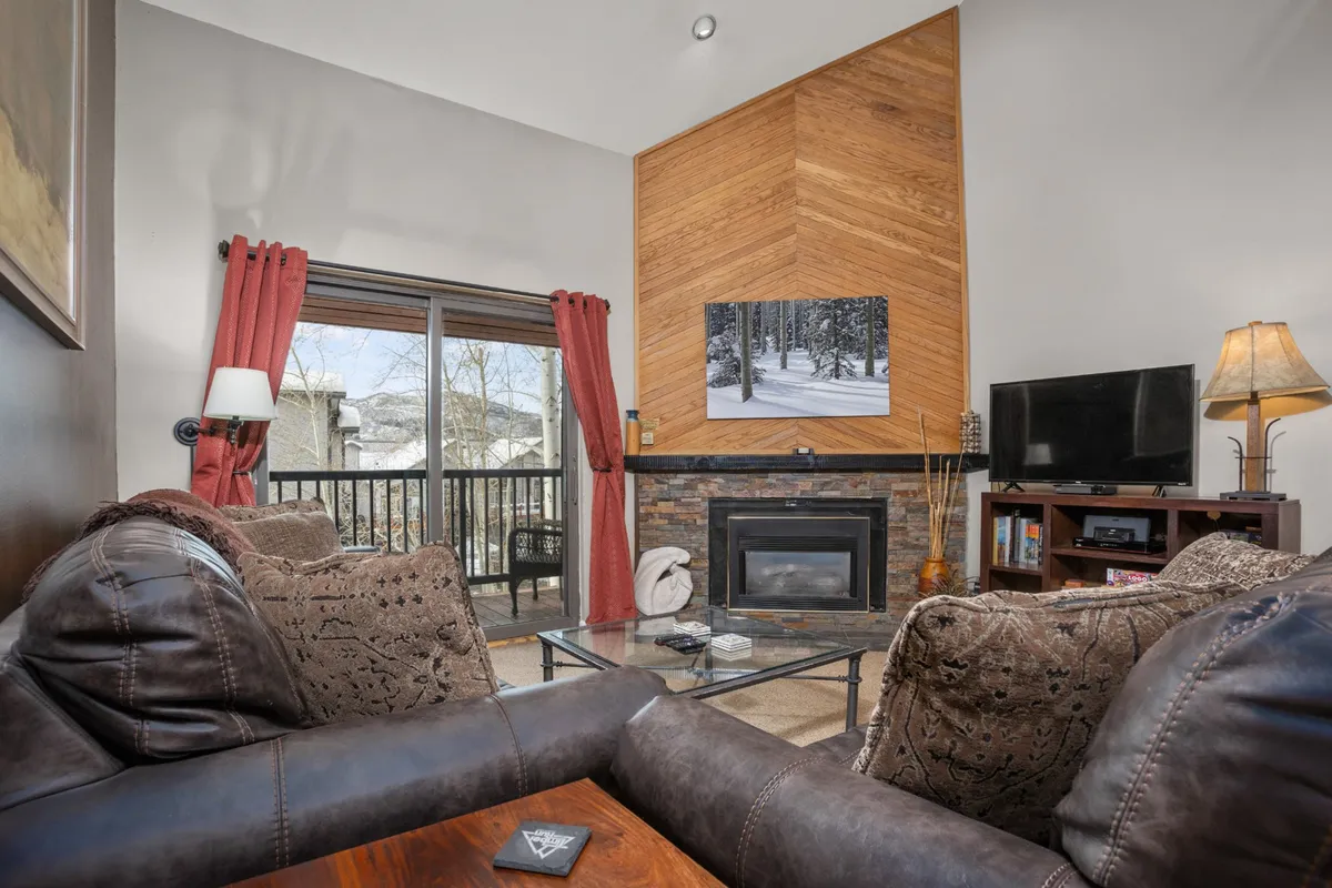 This inviting living area features a prominent stone-surround gas fireplace, plush sofa seating, a large HDTV, and expansive windows that flood the space with natural light and offer views of the winter scenery outside. Wood paneling and warm lighting create a cozy and social atmosphere, perfect for gathering after a day of adventures.