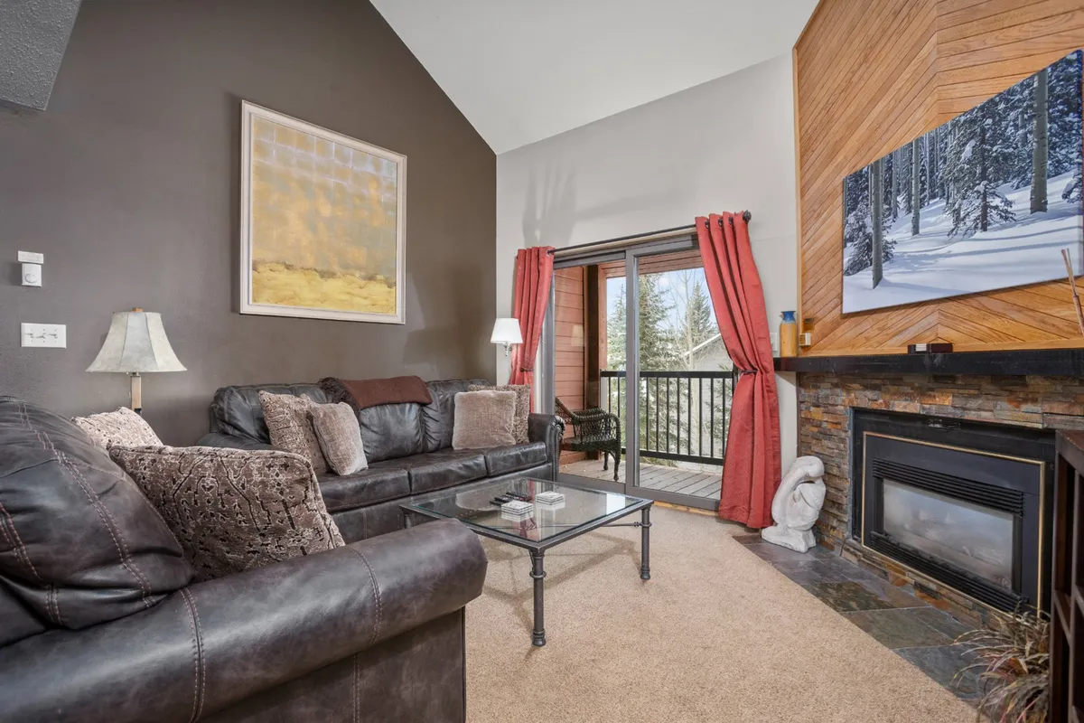 The image showcases a warmly decorated living room with a plush sofa facing a glowing gas fireplace, tasteful mountain-inspired artwork, and large balcony doors letting in natural light and offering a peek at the snowy outdoors. The scene is inviting, with the suggestion of relaxing, socializing, or enjoying a warm drink while watching the snowfall.