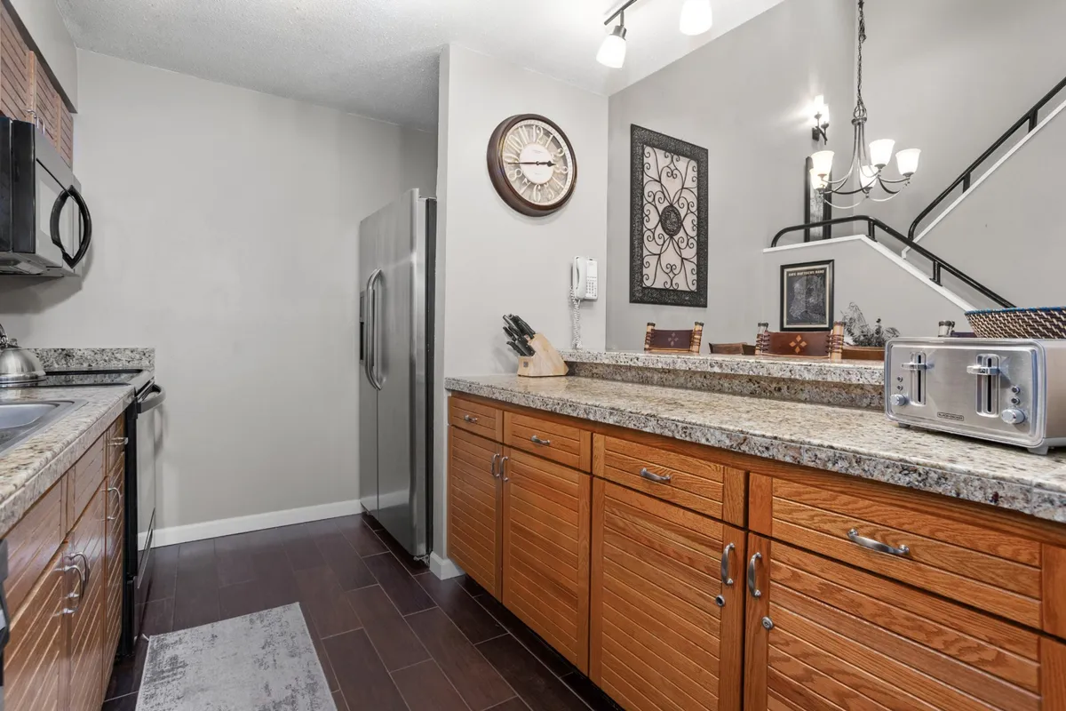 The kitchen features gleaming granite countertops, rich wooden cabinetry, stainless steel appliances, a breakfast bar, and a decorative wall clock. The layout is open and practical, inviting guests to cook, socialize, or grab a quick snack at the bar.