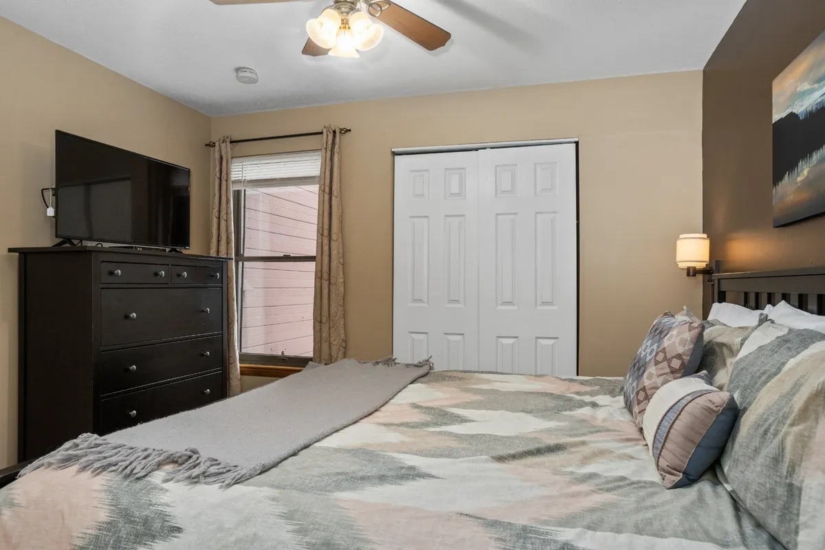 A cozy bedroom featuring a large bed with decorative pillows, a substantial dresser topped with a television, and a window dressed with curtains that let in natural light. The room feels spacious and inviting, ideal for relaxation and unwinding after a day of activity.