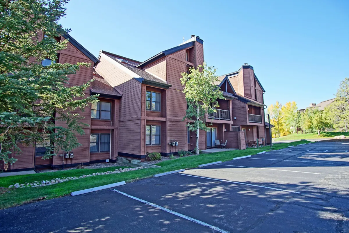 An exterior view showcasing the wooden architecture of Timber Run buildings set among lush greenery and a spacious parking area. The scene suggests easy comings and goings, outdoor gatherings, and quick access to transportation options like the city bus.